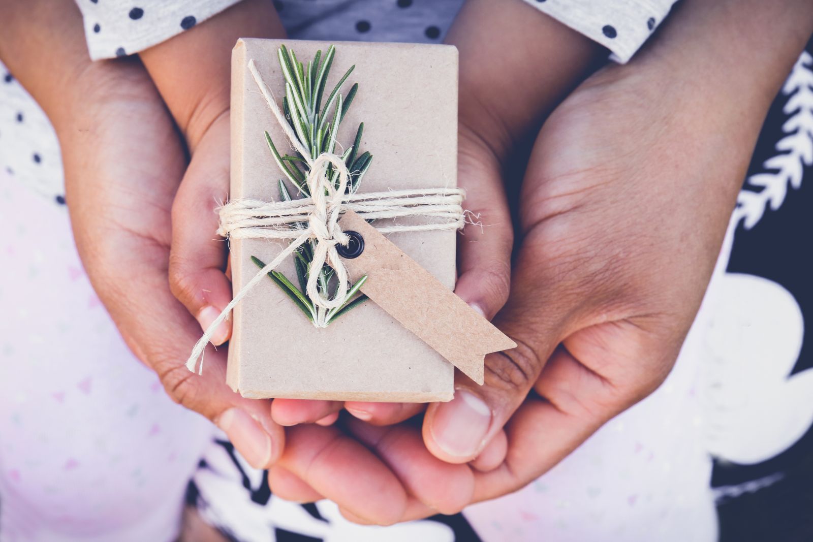 A child's hands are holding a brown paper wrapped box with a sprig of rosemary tucked into the jute ribbon ornament. The child's hands are resting on their parent's cupped hands. The background and the child's sleeves are grey knit fabric with dark blue polka dots.